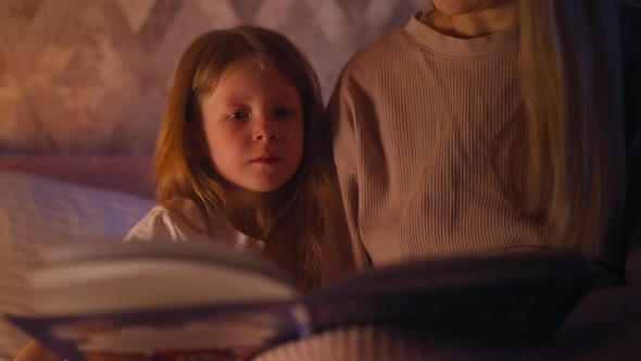 Pretty Child Looks Fairy Tale Book Held By Mother on Bed alt