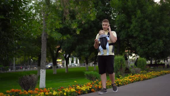A Young Man Walks in the Park with a Newborn Child and Speaks on the Phone alt