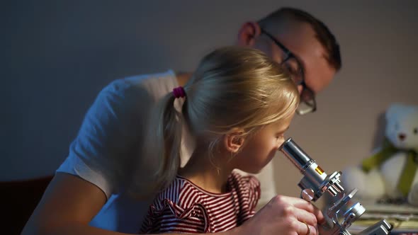 Child Looks Through Microscope While Doing Science Experiments at Home with Her Father
