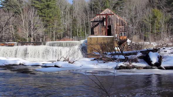 rushing water pours over dam next to dilapidated building. Stationary wide angle alt