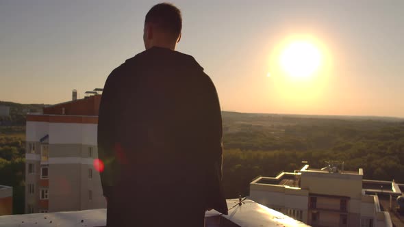 A Man with a Laptop Walks on the Roof at Sunset and Looks at the City From a Height at Sunset alt