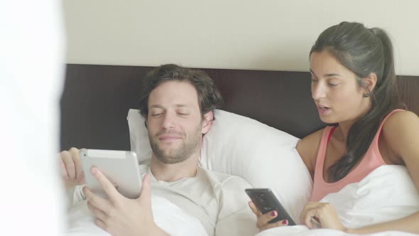 Couple relaxing in bed chatting while using personal wireless devices