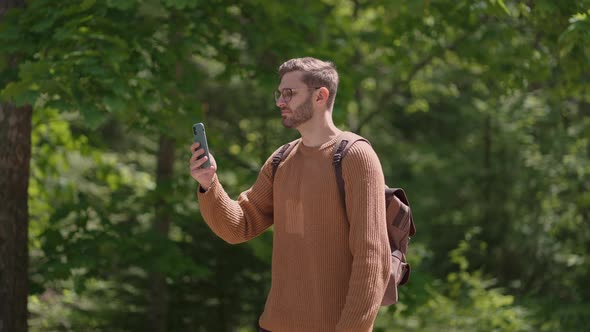 A Young Male Traveler with a Backpack with a Mobile Phone Walks in the Forest Takes Videos alt