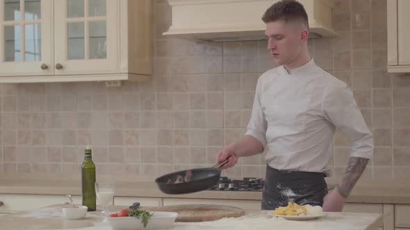 Young Man in Chef Uniform Puts Salt in the Fryng Pan and Throws Up Meat in a Skillet. A Professional alt