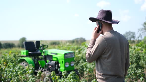 A Young Farmer Uses a Smartphone He is Resting Near His Tractor alt