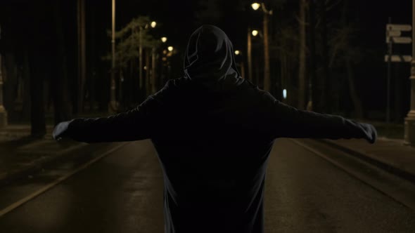 Scary Man Killer or Ghost Stands in Hood on Lit Street at Night and Raises His Hands Up. The Maniac alt