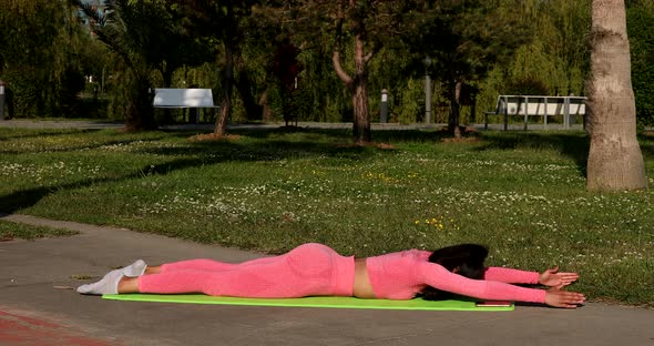 Woman wearing pink sportswear, pants and top, doing stretching yoga exercises on fitness mat in park