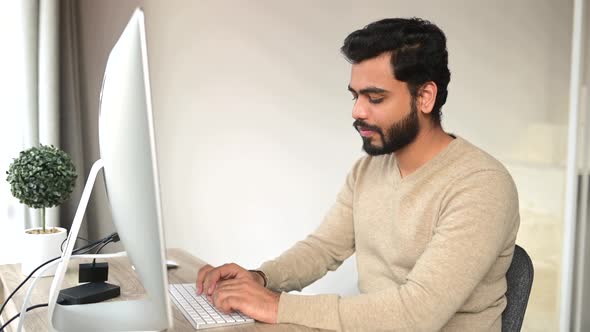 Optimistic Ethnic Man in Casual Wear Using Computer Sitting at the Table