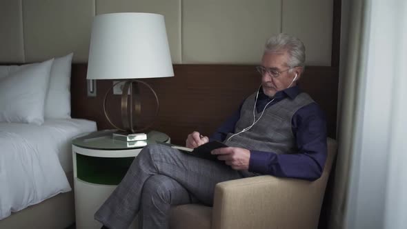 Portrait of an Grayhaired Designer Sitting in a Hotel Room and Writing Down Thoughts in a Notebook alt