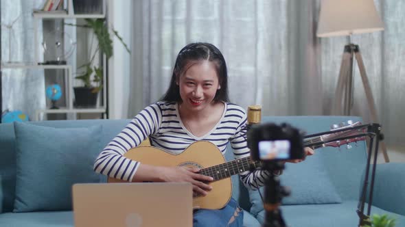 Woman With A Laptop Waving Hand And Speaking To Camera While Live Stream Playing A Guitar At Home alt