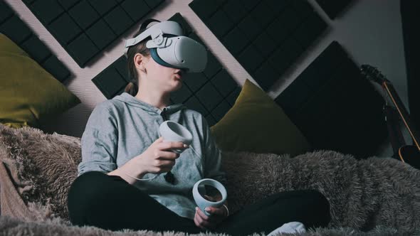 Young Woman Sitting on Sofa Playing Game Using VR Helmet Indoor at Home alt
