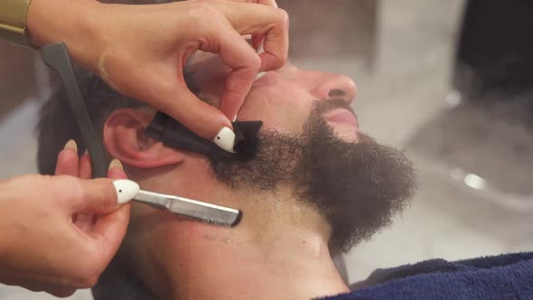 Barber Shaving Bearded Man with Sharp Razor in Barber Shop