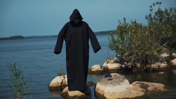 Woman devil in black cape and hood on stones near the river. Halloween costume. alt