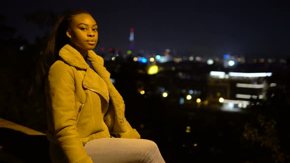 A Young Black Woman Looks Around and at the Camera As She Sits on a Low Wall in Urban Area at Night alt
