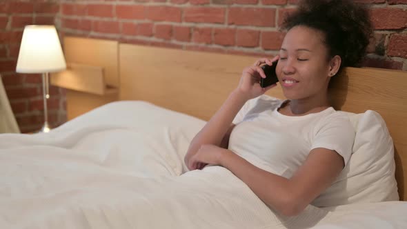 African Woman Talking on Call on Smartphone in Bed alt