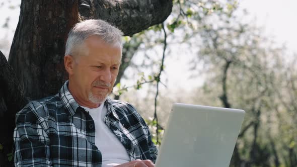 An Elderly Whitehaired Male Businessman with a Beard Uses a Laptop on the Street in a Park on a