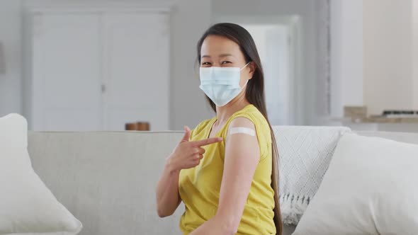Happy asian woman in face mask sitting on sofa showing arm with plaster after vaccination alt