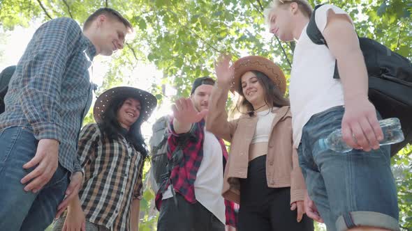 Bottom Angle View of Positive Caucasian Young Men and Women Stacking Hands in Sunlight Outdoors alt