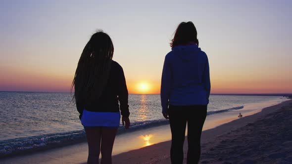 Two Women Hold Hands on a Background of Sunset and Sea Waves alt
