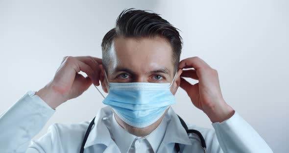 Portrait of Male Doctor Putting on Medical Mask and Looks at the Camera Over White Background alt