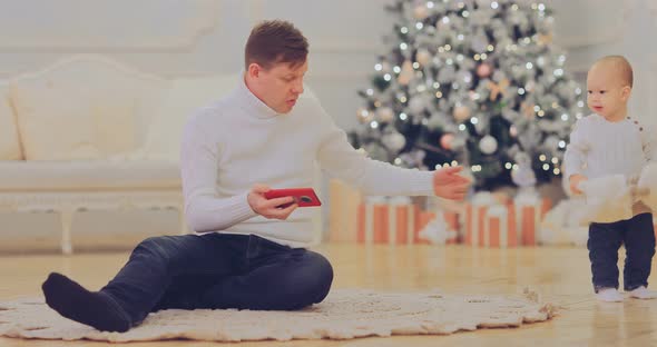 Dad Shows Pictures on the Phone Sits on the Floor By the New Year Tree at Home