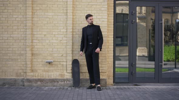 Wide Shot Portrait of Confident Caucasian Businessman Standing on City Street with Skateboard alt
