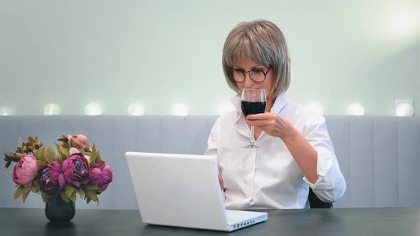 A Happy Beautiful Elderly Lady Drinking Wine Calls a Friend a Relative From Home on a Laptop Video