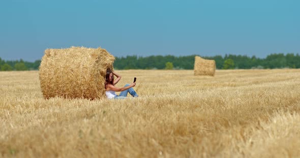 Happy Woman is Sitting in a Field By a Haystack and Taking a Selfie on Her Mobile Phone alt