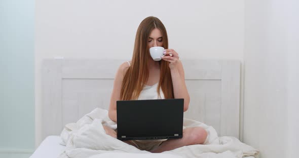 Young Woman Freelancer Is Sitting on the Bed Drinking Coffee and Working on a Laptop, Morning Time alt