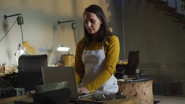 Caucasian female jeweller in workshop wearing apron, sitting at desk, using laptop alt