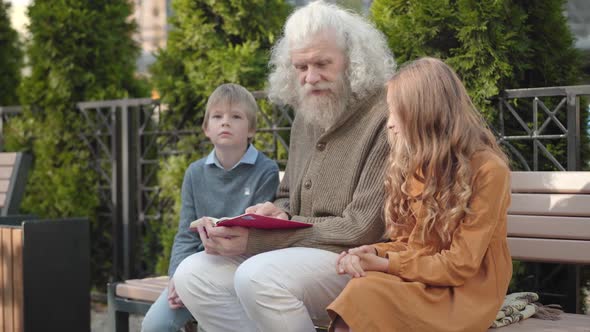 Angle View of Intelligent Male Retiree Explaining Book To Grandchildren on Sunny Day Outdoors alt