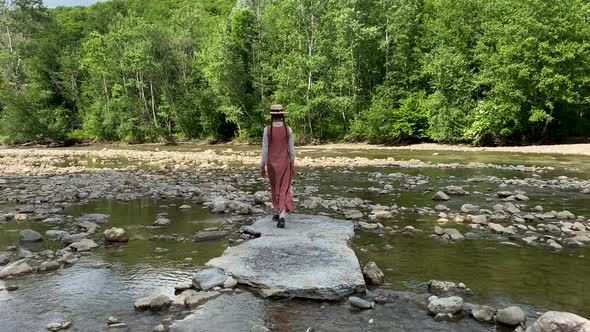 Vintage romantic woman in straw hat, long dress walks on river rocks near forest. Natural scenic