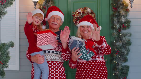 Senior Grandmother Grandfather with Granddaughter Standing at Christmas House Porch Waving Hello Hi alt