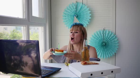 A woman blows out a candle on a cake and looks into a laptop.