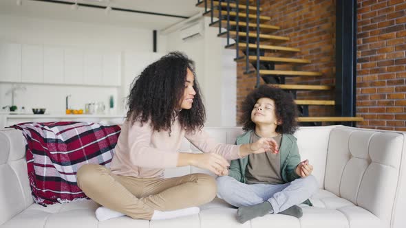 Curlyhaired Mother and Son are Sitting on the Sofa in the Lotus Position alt