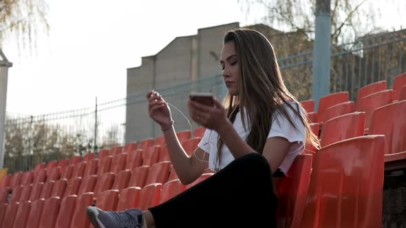 Young Fit Asian Woman with Headphones Listens to Music on Smartphone at Stadium