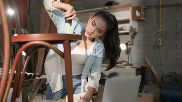 Young Asian designer furniture woman Carpenter uses a tape measure to measure chair on workbench