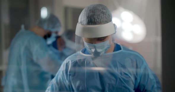 Portrait of Male Surgeon Standing Near Glass Door While Team Performing Surgery in Operation Room alt