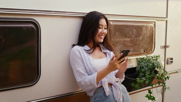 Smiling Young Asian Woman Using Smartphone While Standing Outdoors Near Modern Trailer alt