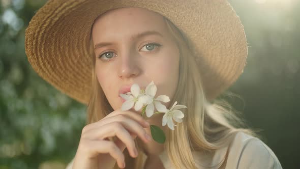 Portrait of Blonde Woman in Hat Squeezing Petals of White Apple Blossoms alt
