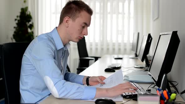 Young Handsome Man Works on Desktop Computer and Reads Document in the Office alt