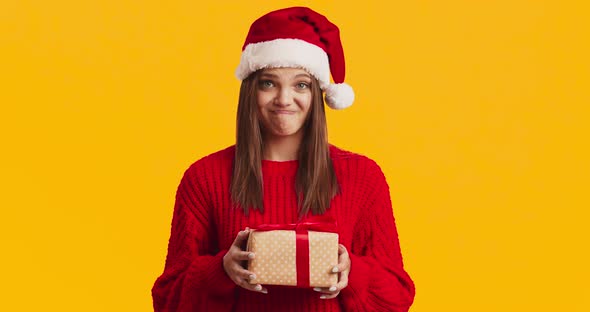 Young Displeased Woman in Santa Hat Holding Present Box and Frowning, Orange Studio Background alt