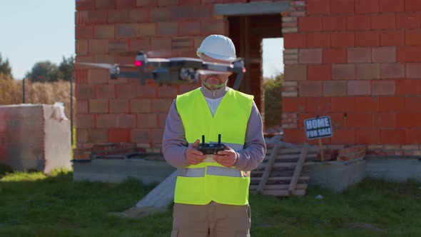 Drone Operator Holding Remote Controller Controls an Aircraft in Front of Construction House Site alt