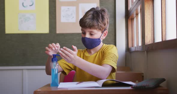 Boy wearing face mask sanitizing his hands while sitting on his desk at school alt