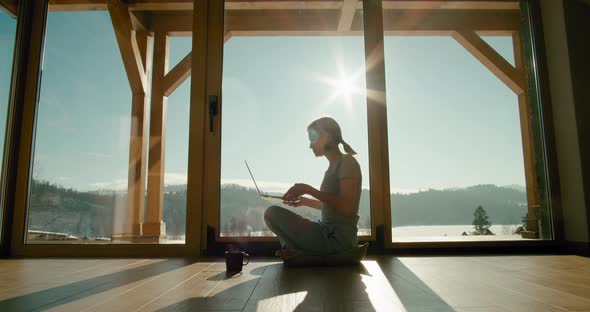 Young Woman Uses Laptop Sitting on Floor By Big Window with View on Mountains