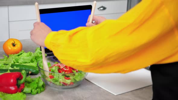 Woman in Kitchen Mixes Cooked Vegetarian Salad with Wooden Spoons in Glass Bowl alt