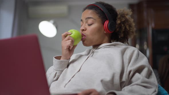 Confident African American Woman in Headphones Eating Apple Messaging Online on Laptop alt