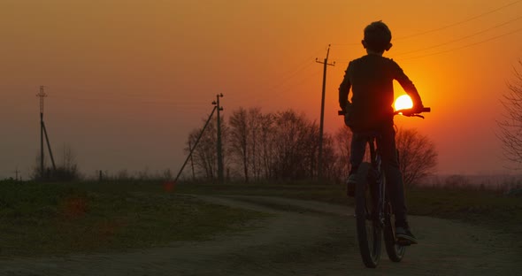 A Boy Is Riding a Bicycle on a Pathway at Sunset. Filming From the Back.  alt
