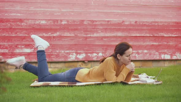 A girl with a laptop is lying on a blanket on the lawn and communicating via video link alt