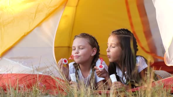 Two Little Girls are Sisters They are Sitting in a Tent and Blowing Bubbles alt
